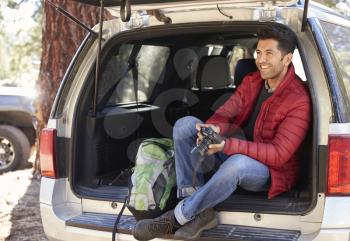 Man relaxing in open back of car holding camera