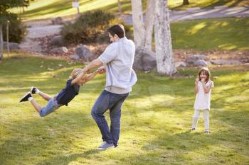 Father Swinging Son By His Arms In Park As Daughter Watches