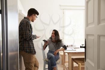 Two teenage friends using smartphones, talking in kitchen