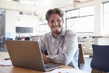 Young man in office using laptop computer smiling to camera