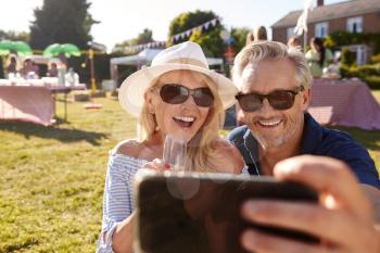 Mature Couple Sitting On Rug At Summer Garden Fete Taking Selfie On Mobile Phone