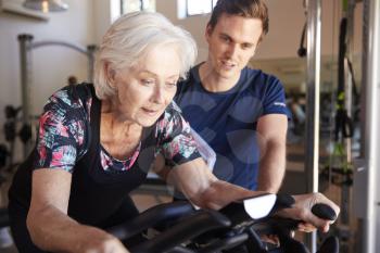 Senior Woman Exercising On Cycling Machine Being Encouraged By Male Personal Trainer In Gym