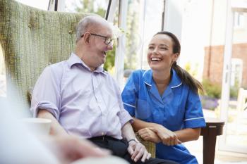 Senior Man Sitting In Chair And Talking With Nurse In Retirement Home