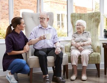 Male And Female Residents Sitting In Chair And Talking With Carer In Retirement Home