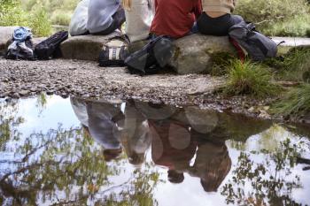Reflection in stream of five young adult friends taking a break sitting on rocks during a hike, back view, low section