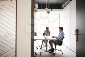 Businessman And Businesswoman Having Discussion Around Boardroom Table Viewed Through Meeting Room Door