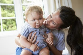 Mother Hugging Happy Baby Son By Window At Home