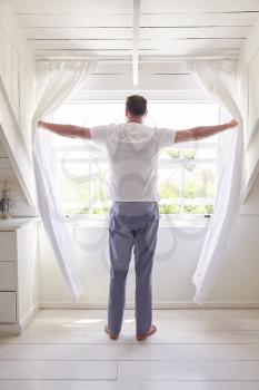 Rear View Of Man Opening Curtains And Looking Out Of Window
