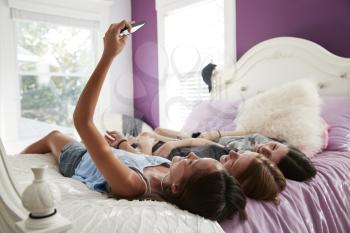 Teenage girl taking a selfie with two friends lying on a bed