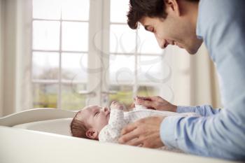 Father Playing With Newborn Baby Lying On Changing Table