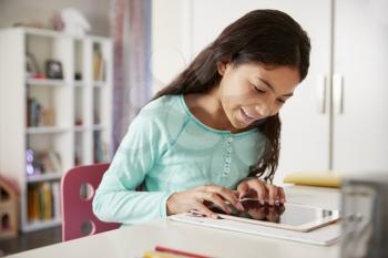 Young Girl Sitting At Desk In Bedroom Using Digital Tablet To Do Homework