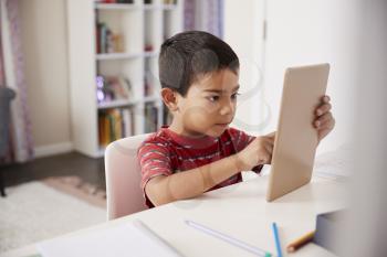 Young Boy Sitting At Desk In Bedroom Using Digital Tablet To Do Homework