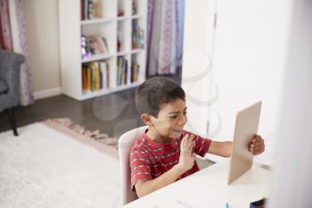 Young Boy Sitting At Desk In Bedroom Using Digital Tablet To Make Video Call
