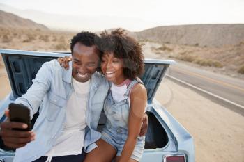 Couple Posing For Selfie Sitting In Trunk Of Classic Car On Road Trip