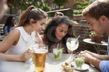 Friends Sitting At Table In Pub Garden Looking At Message On Mobile Phone