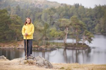 A pre-teen girl standing on a rock holding a stick, smiling to camera, lakeside background
