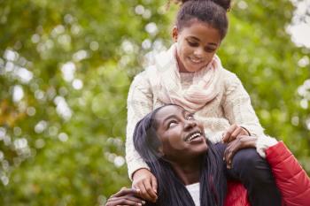 Young black woman carrying her pre-teen daughter on her shoulders in the park, both smiling at each other, close up