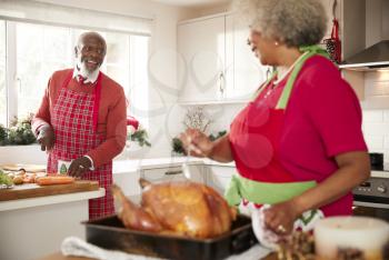 Senior black woman preparing a roast turkey for Christmas dinner turns to talk to her husband, chopping vegetables in the background, close up
