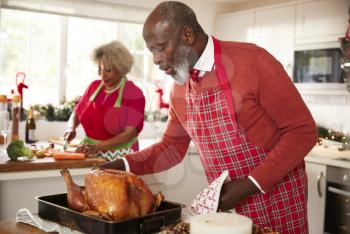 Senior black man basting roast turkey in preparation for Christmas dinner, his wife chopping vegetables in the background, close up