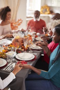 Multi generation mixed race family holding hands and saying grace before eating at their Thanksgiving dinner table, selective focus, vertical