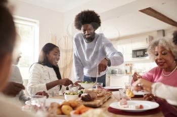 Middle aged black man carving and serving meat at Sunday family dinner with his partner, kids and their grandparents, front view