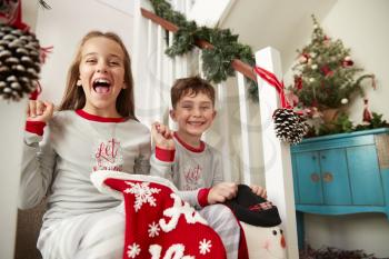 Portrait Of Two Excited Children Wearing Pajamas Sitting On Stairs Holding Stockings On Christmas Morning