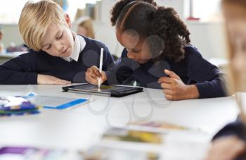 Schoolgirl using a tablet and stylus sitting with a boy at a desk in a primary school classroom, selective focus