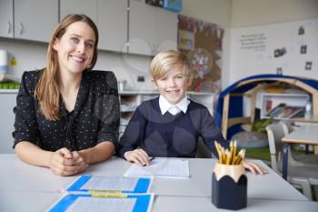 Young female primary school teacher and schoolboy sitting at a table, working one on one in a classroom, smiling to camera, front view, close up