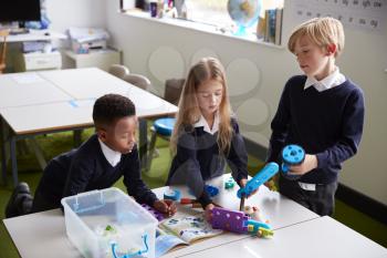 Elevated view of three primary school kids standing at a table in a classroom, working together with toy construction blocks