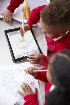 Elevated view of two kindergarten school kids sitting at a desk in a classroom drawing with a tablet computer and stylus, close up, vertical