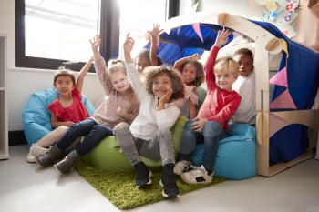 A multi-ethnic group of infant school children sitting on bean bags in a comfortable corner of the classroom, raising their hands to answer a question, low angle, close up