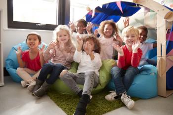 A multi-ethnic group of infant school children sitting on bean bags in a comfortable corner of the classroom, smiling and waving to camera, low angle, close up