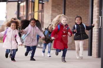 Happy young school girls wearing coats and carrying schoolbags running in a walkway with their classmates outside their infant school building