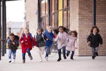 A happy multi-ethnic group of young school kids wearing coats and carrying schoolbags running in a walkway with their classmates outside their infant school building