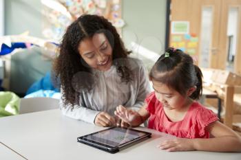 Female infant school teacher working one on one with a Chinese schoolgirl, sitting at a table in a classroom using a tablet computer, close up