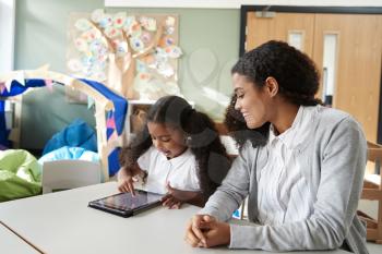 Young black schoolgirl sitting at a table in an infant school classroom using a tablet computer and learning one on one with a female teacher, close up
