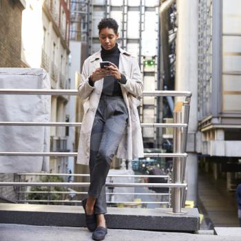 Fashionable young black woman standing in the city leaning on a hand rail using her smartphone, full length, low angle