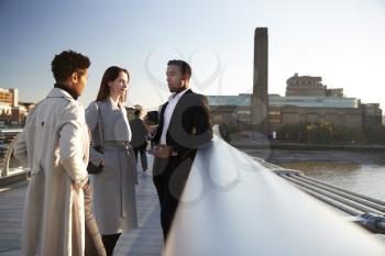 Side view of three millennial business colleagues stand talking on Millennium Bridge, London, selective focus