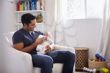 Smiling Hispanic father holding his four month old son feeds him a bottle at home