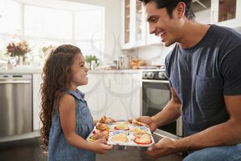 Smiling young Hispanic girl standing in kitchen presenting the cakes she has baked to her father