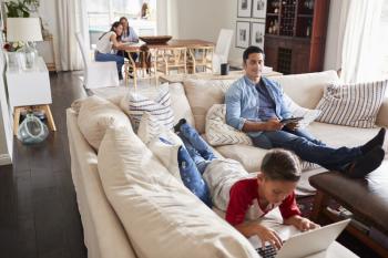 Pre-teen boy lying on sofa using laptop, dad sitting with tablet, mum and grandma in the background