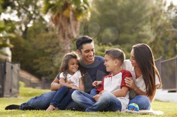 Happy young Hispanic family sitting together on the grass in the park, looking at each other