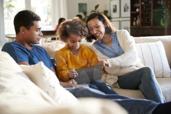 Young parents and their pre-teen daughter sitting on a sofa in the living room using a tablet computer together, selective focus