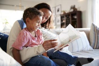 Close up of young mother sitting on a sofa in the living room reading a book with her toddler son, who is sitting on her knee, side view
