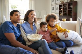 Young family sitting together on the sofa in their living room watching TV and eating popcorn