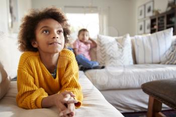 Close up of pre-teen girl lying on sofa watching TV in the living room, her younger brother sitting in the background, focus on foreground