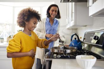 Pre-teen girl standing at hob in the kitchen using spatula and frying pan, preparing food with her mother, side view