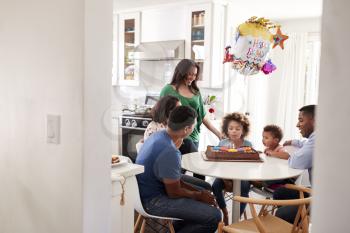 Pre teen girl sitting at the kitchen table with her three generation family celebrating her birthday, blowing out candles on her birthday cake, seen from doorway