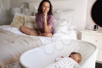 Young adult mother sitting cross legged on her bed looking down at her three month old baby sleeping in his cot, close up