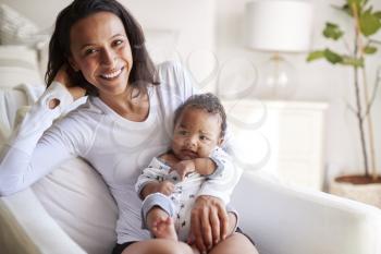 Happy mixed race young adult mother sitting on an armchair holding her three month old baby son, smiling to camera, close up